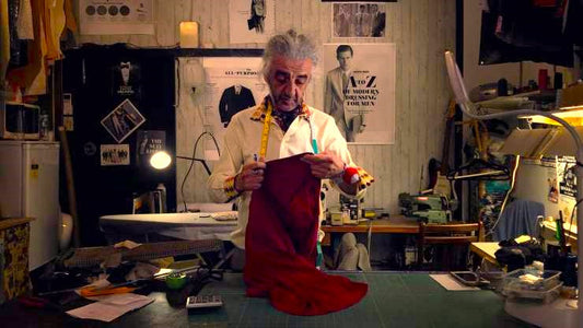 Ferudun Avar aka West End Tailor tailor in a vintage sewing workshop examining a piece of red fabric under warm lighting, surrounded by sewing tools, patterns, and posters on the wall.