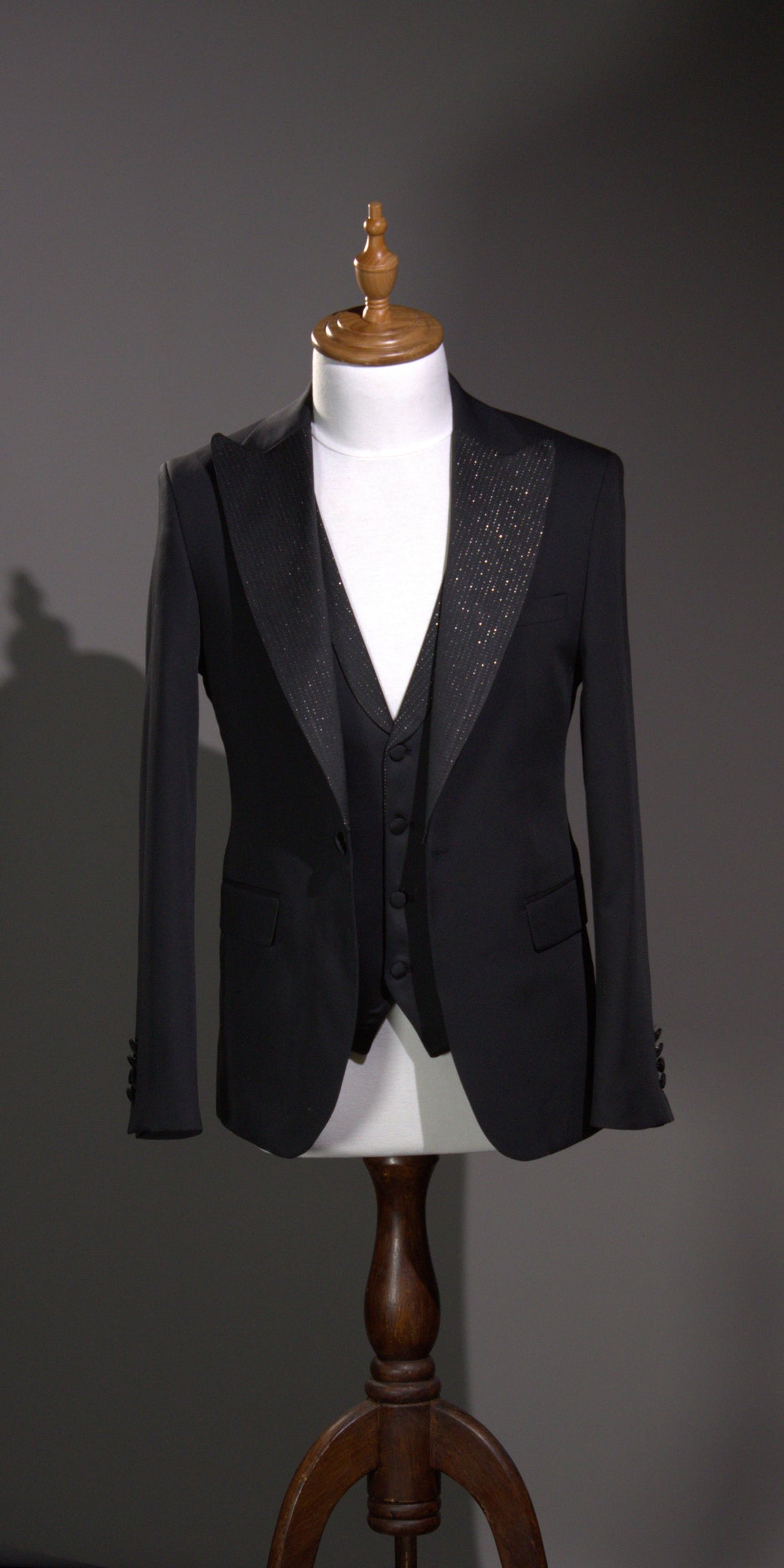 Tailored black dinner tuxedo jacket with black satin peak lapels and a matching low-cut black vest on a tailor mannequin.
